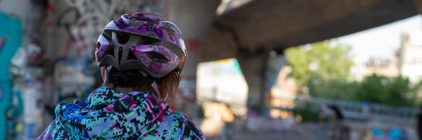 A helmeted bicyclist (me) looks towards an out-of-focus skate park built around bridge supports.