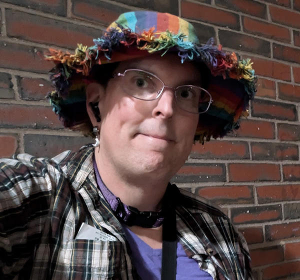 A white woman in a colorful, fringed hat, smirking/smiling at the camera.